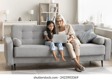 Positive smart grandma teaching child to use application on laptop, helping with school homework tasks, sitting on cozy sofa with computer on lap in modern home interior - Powered by Shutterstock