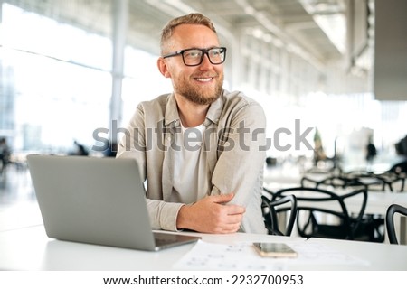 Positive smart successful caucasian man, with glasses, in stylish casual wear, IT specialist, company seo, freelancer, sits at a desk with a laptop in coworking, looks away, smiles happily
