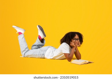 Positive smart teen black curly girl in white t-shirt, glasses reads book, lies on floor, isolated on yellow studio background. Study, knowledge, hobby, rest and relax, ad and offer - Powered by Shutterstock
