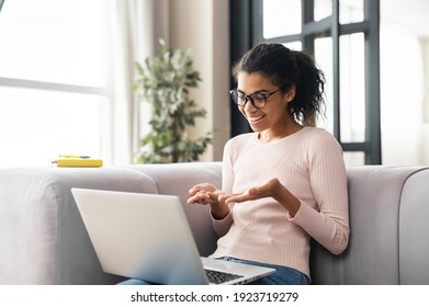 Positive smart young African American female freelancer or a student in glasses sitting on the couch with laptop on the lap, studying from online webinar or a course at home, video calling and talking