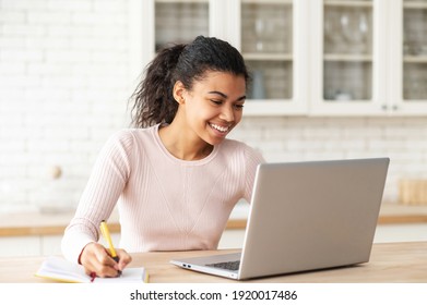 Positive Smart Young Mixed-race Female Student Smiling Sitting At The Table With Laptop In The Kitchen, Studying From Home And Taking Notes To The Notebook, Watching An Online Webinar Or A Course