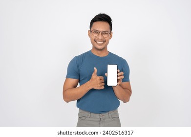A positive, smiling Asian man in casual clothes is showing his thumb up and a white-screen smartphone mockup to the camera, standing on an isolated white background. People and technology concepts