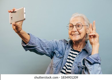 Positive smiling elderly woman wearing casual clothes and glasses poses for selfie holding smartphone in one hand and making peace or victory sign with another. Active old age concept