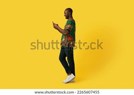Positive smiling happy mature african american man in traditional costume using modern cell phone over yellow studio background, checking newest mobile app, copy space, full length shot