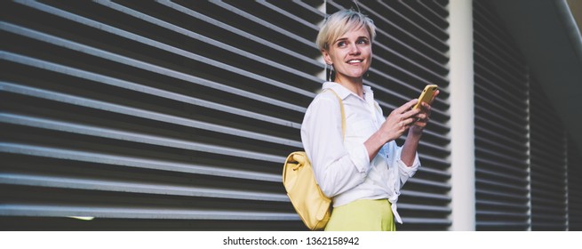 Positive Smiling Hipster Girl With Cellular Phone In Hand Looking Away While Waiting Friend On Urban Setting, Happy Millennial Female Blogger Using Free Wifi On Publicity Area For Chatting Online