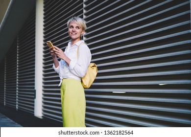 Positive Smiling Hipster Girl With Cellular Phone In Hand Looking Away While Waiting Friend On Urban Setting, Happy Millennial Female Blogger Using Free Wifi On Publicity Area For Chatting Online