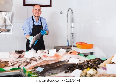 Positive smiling mature salesman with apron offering fresh fish in shop