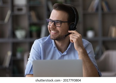 Positive smiling millennial male wear headphones with mic call center worker telephonist operator look aside of laptop screen. Happy young guy remote consultant freelancer feel glad helping customers