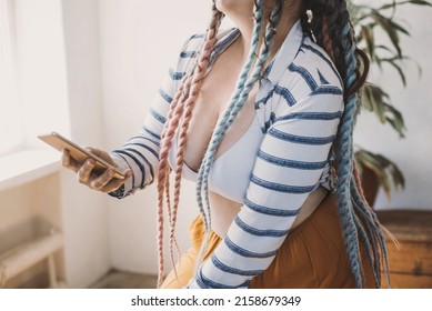 Positive Smiling Plus Size Woman With Dreadlocks In Bright Yellow Pants In A Modern Bohemian Boho Interior Using Smartphone. Online Shopping At Home. No Face