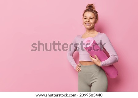 Positive smiling slim woman in tracksuit poses with rolled karemat does yoga pilates or fitness exercises has healty body isolated over pink background blank space aside for your promotional content