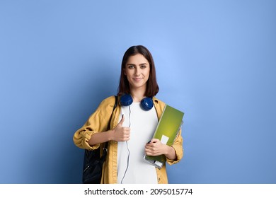 Positive Smiling Student Girl Is Showing Thumb Up, Likes An Idea, Supports Initiative, Ready To Join The Group, Holding Books, Wearing Yellow Shirt, White T-shirt, Black Bag And Headphones Over Neck