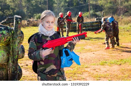 Positive Smiling Teen Girl Wearing Uniform And Holding Gun Ready For Playing With Friends On Paintball Outdoor