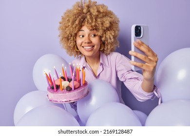 Positive smiling woman celebrates special occasion poses for selfie to share photo with friends holds big tasty cake surrounded by inflated balloons isolated over purple background. Holiday concept - Powered by Shutterstock