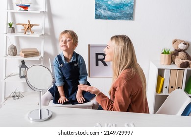 Positive Speech Therapist Talking And Holding Clipboard Near Child And Mirror In Consulting Room