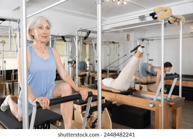 Positive sporty aged woman doing remedial gymnastic exercises on Pilates reformer to improve joints mobility and overall strength in modern fitness center - Powered by Shutterstock