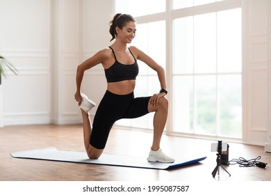 Positive Sporty Lady Stretching Leg In Front Of Smartphone On Tripod At Home, Free Copy Space. Beautiful Female Fitness Blogger Recording Video For Her Followers Or Having Personal Training Online