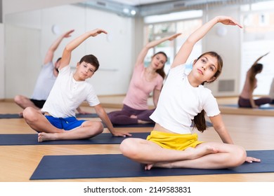 Positive Sporty Preteen Girl Doing Stretching Exercises While Sitting In Lotus Position During Training With Family In Fitness Center