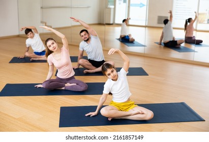 Positive Sporty Preteen Girl Doing Stretching Exercises While Sitting In Lotus Position During Training With Family In Fitness Center