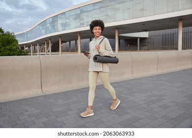 Positive sporty woman returns from sport training carries rolled kareamt uses mobile phone for chatting online wears hoodie leggings and sneakers has happy mood after successful outdooe workout - Powered by Shutterstock