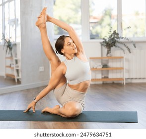 Positive sporty young woman demonstrating harmonious strength and flexibility of seasoned yogini performing advanced yoga posture on mat in serene studio with natural light.. - Powered by Shutterstock