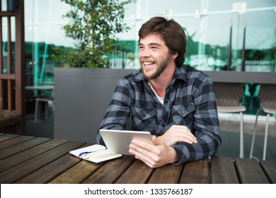Positive student enjoying wireless connection in street cafe. Joyful guy in casual sitting at table outdoors, holding tablet and laughing at funny scene. Communication concept