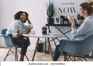 Positive Stylish Dark Skinned Girl Enjoying Tasty Coffee And Good Communication Together With Best Friend At Home With Cool Interior.Two Cheerful Women Talking About Funny Stories Sitting At Table