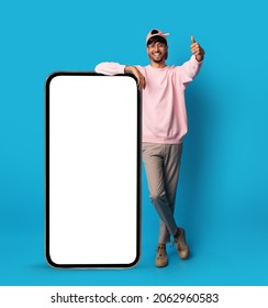 Positive stylish indian guy posing with big smartphone with empty screen, showing thumb up and smiling, recommending newest mobile app, blue studio background, mockup, full length shot - Shutterstock ID 2062960583
