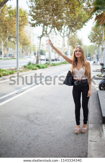 Positive stylish lady waving hand while catching\
taxi outdoors. Content attractive young woman waiting for taxi in\
street. City life\
concept
