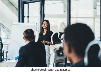 Positive Successful Female Trader Talking About Financial Reports During Briefing In Office.Cheerful Young Woman Coach Conducting Business Workshop For Young Entrepreneurs Standing Near Flip Chart