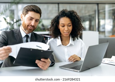Positive successful work colleagues, a caucasian man and an african american woman, are sitting in office, working on a laptop and with documents, discussing and analyzing data, compiling reports