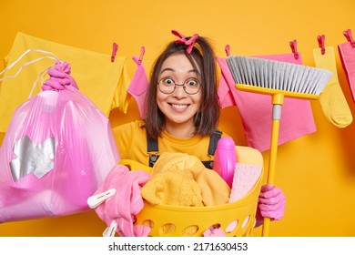 Positive Surprirsed Asian Housewife Holds Plastic Bag Of Litter And Broom Does Household Chores Wears Spectacles Rubber Gloves Poses Near Basket Full Of Laundry Maintains Housekeeping On Weekend