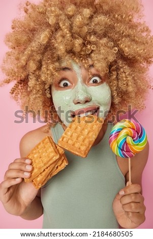 Positive surprised woman applies nourishing clay mask eats appetizing waffles and multicolored lollipop dressed in t shirt isolated over pink background. Facial treatments and pampering concept