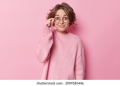 Positive Surprised Woman Keeps Hand On Rim Of Spectacles Smiles Pleasantly Reacts On Good News Wears Casual Cashmere Jumper Hears Something Awesome Isolated Over Pink Background. Reaction And Emotions