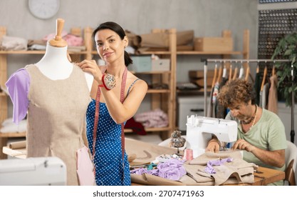 Positive tailoress working with dummy while middle-aged tailor using sewing-machine in workshop