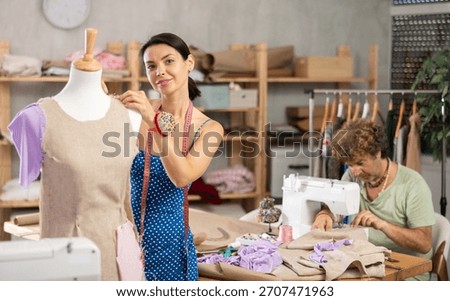 Positive tailoress working with dummy while middle-aged tailor using sewing-machine in workshop