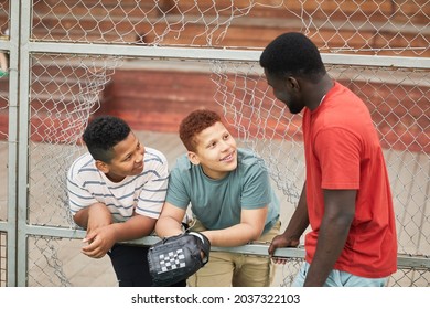 Positive teenage boys leaning on plank of metal net fence with hole and chatting with elder brother playing baseball - Powered by Shutterstock