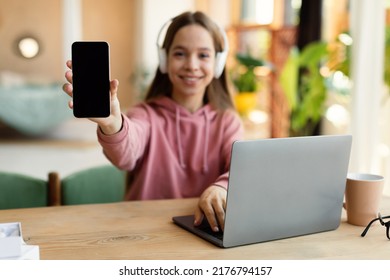 Positive Teenage Lady Demonstrating Blank Smartphone While Studying At Desk With Laptop At Home, Mockup, Free Space. Female Student Advertising Educational App Or Mobile Website