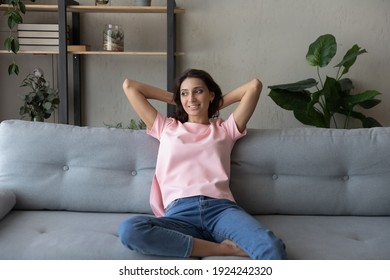 Positive thinking. Happy young indian woman relax on couch at cozy apartment hold hands behind head look aside. Tranquil arab female taking break from work daydreaming enjoying silence peace at home