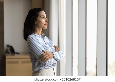 Positive thoughtful confident Latin business owner woman standing with arms crossed in office at large window, looking away in deep thoughts, thinking on job success, successful project plan - Powered by Shutterstock