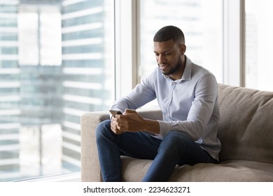 Positive thoughtful young African business man using mobile phone for online communication, sitting on couch in modern home flat with big window glass, typing, chatting on Internet - Shutterstock ID 2234623271