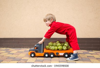 Positive Toddler Boy In A Red Overalls Carries A Lot Of Organic Farm Apples On A Big Toy Car - A Truck. Courier, Truck Driver, Like Dad. Games For Boys