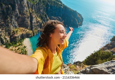 Positive tourist woman taking picture outdoors for memories, making selfie on top of cliff with valley mountains view, sharing travel adventure journey - Shutterstock ID 2116336466