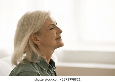 Positive tranquil blonde senior woman meditating at home, sitting with closed eyes, keeping silence in deep thoughts, breathing fresh air, smiling, enjoying relaxation, break. Side view - Powered by Shutterstock
