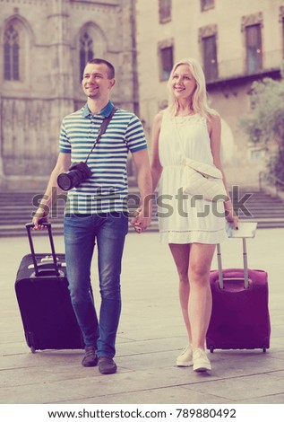 Positive traveling man and woman walking together with luggage and looking around in city