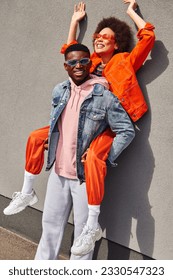 Positive and trendy young african american man in denim jacket holding best friend in sunglasses and bright outfit and standing near building on urban street, trendy friends in urban settings - Shutterstock ID 2330547323