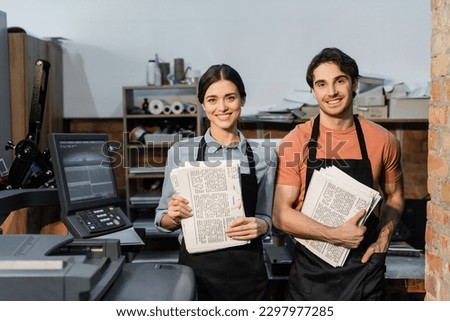 positive typographers in aprons smiling while holding printed newspapers
