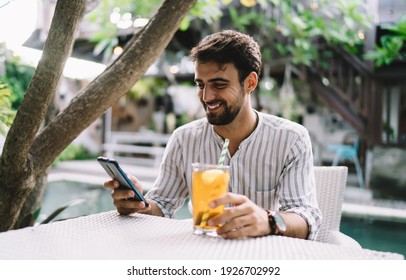 Positive unshaven male in casual wear sitting at table with glass of colorful refreshing drink while surfing internet on cellphone in cafe near tree trunk - Shutterstock ID 1926702992