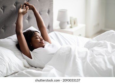 Positive well-rested african american millennial woman stretching in comfy bed, stretching hands and smiling with closed eyes, enjoying new day, wearing pajams, panorama with copy space