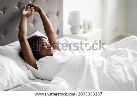 Positive well-rested african american millennial woman stretching in comfy bed, stretching hands and smiling with closed eyes, enjoying new day, wearing pajams, panorama with copy space