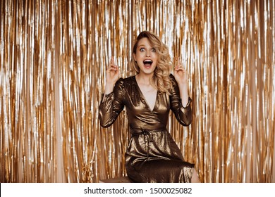 Positive woman in brilliant dress emotionally and surprised looks into camera. Blonde crosses fingers on gold background